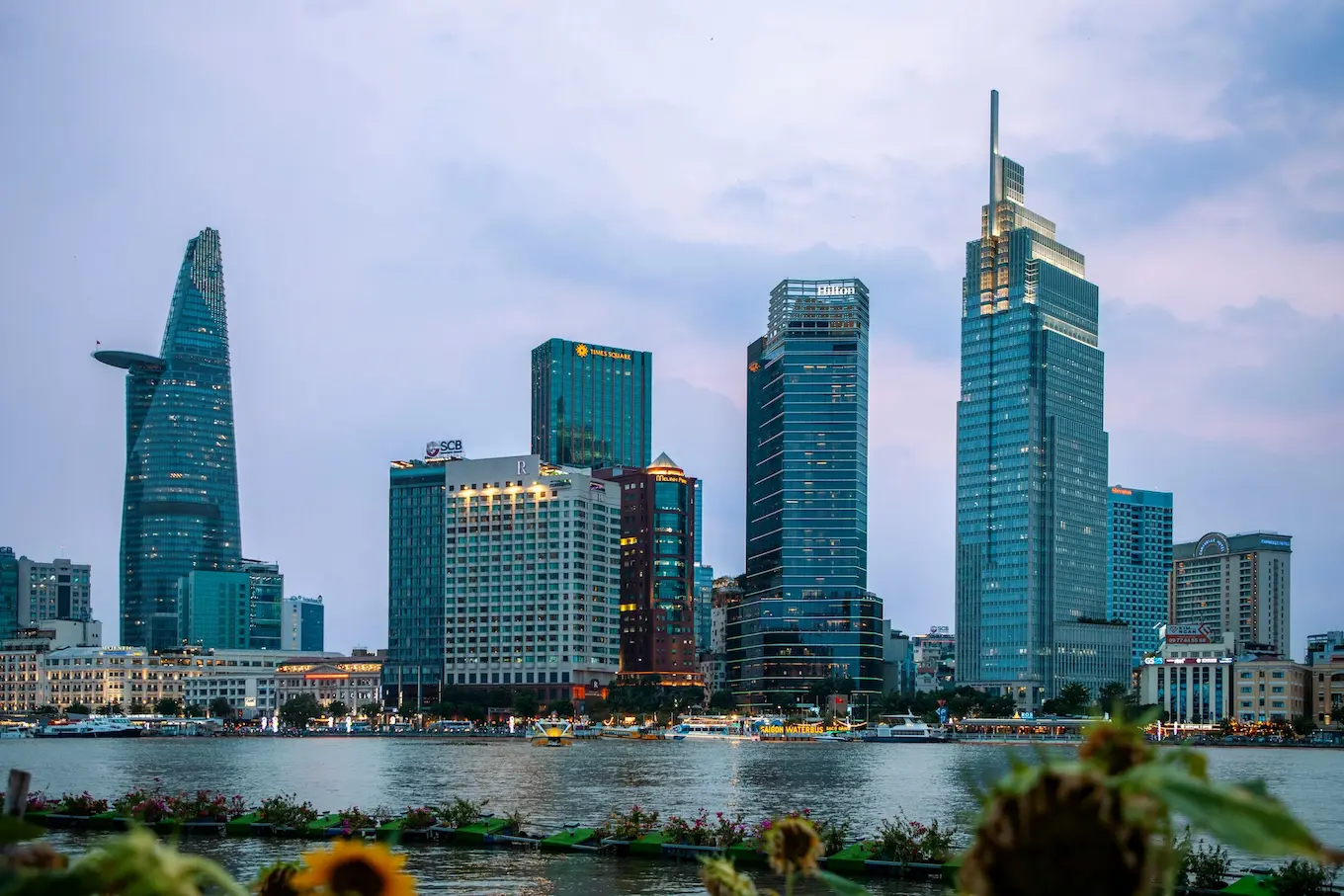 Skyline of a city at dusk, featuring tall skyscrapers and a river in the foreground, with lights reflecting on the water.