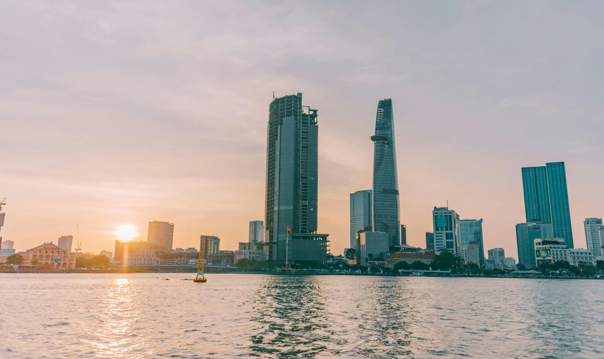 City skyline at sunset with tall buildings by a river, reflecting warm sunlight on the water.