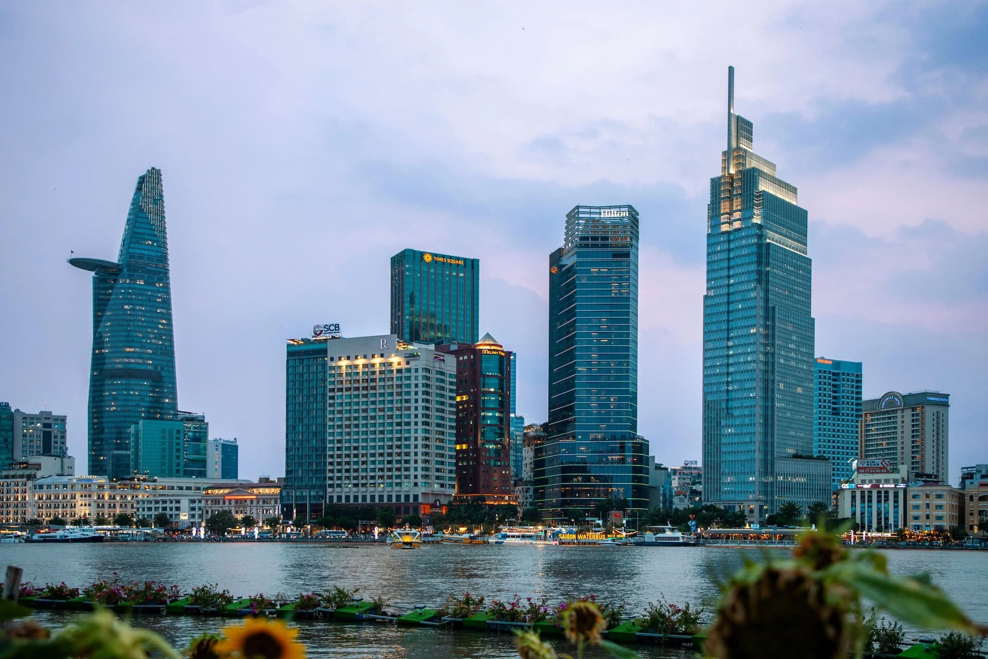 Skyline of a city with modern skyscrapers by a river, under a cloudy sky. Sunflowers are visible in the foreground.